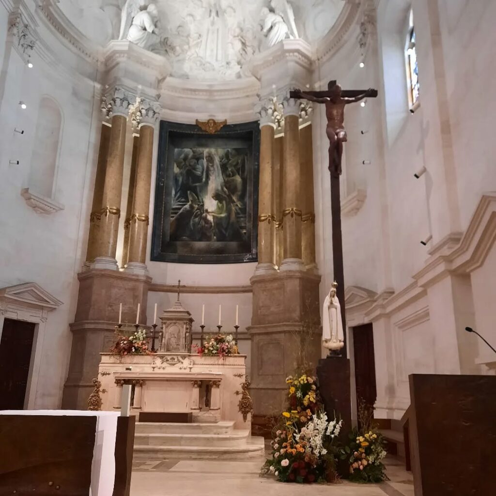 Crucifixo em madeira e escultura de Nossa Senhora em primeiro plano, com o altar e retábulo ao fundo, em capela do Santuário de Fátima.