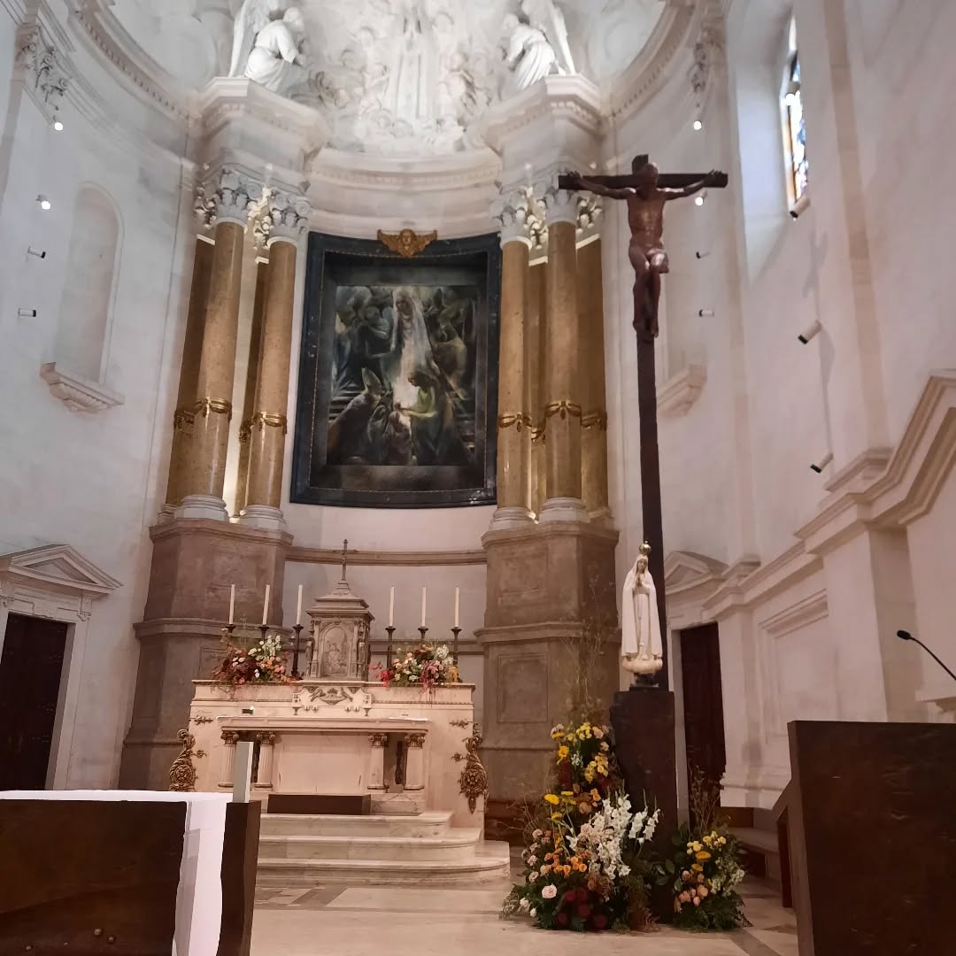 Crucifixo em madeira e escultura de Nossa Senhora em primeiro plano, com o altar e retábulo ao fundo, em capela do Santuário de Fátima.