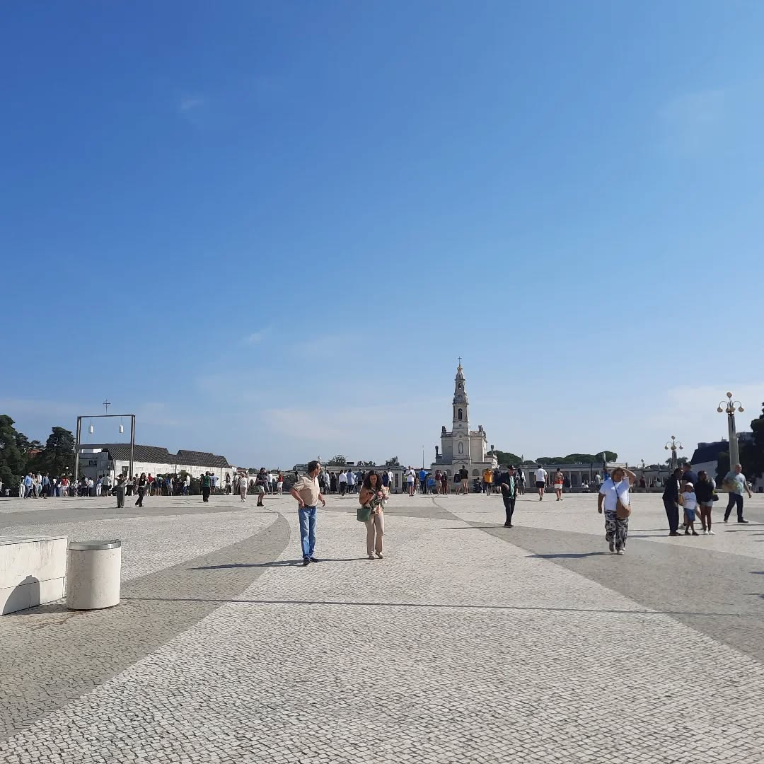 Peregrinos caminhando sob céu azul na esplanada do Santuário de Fátima, com a Basílica do Rosário ao fundo
