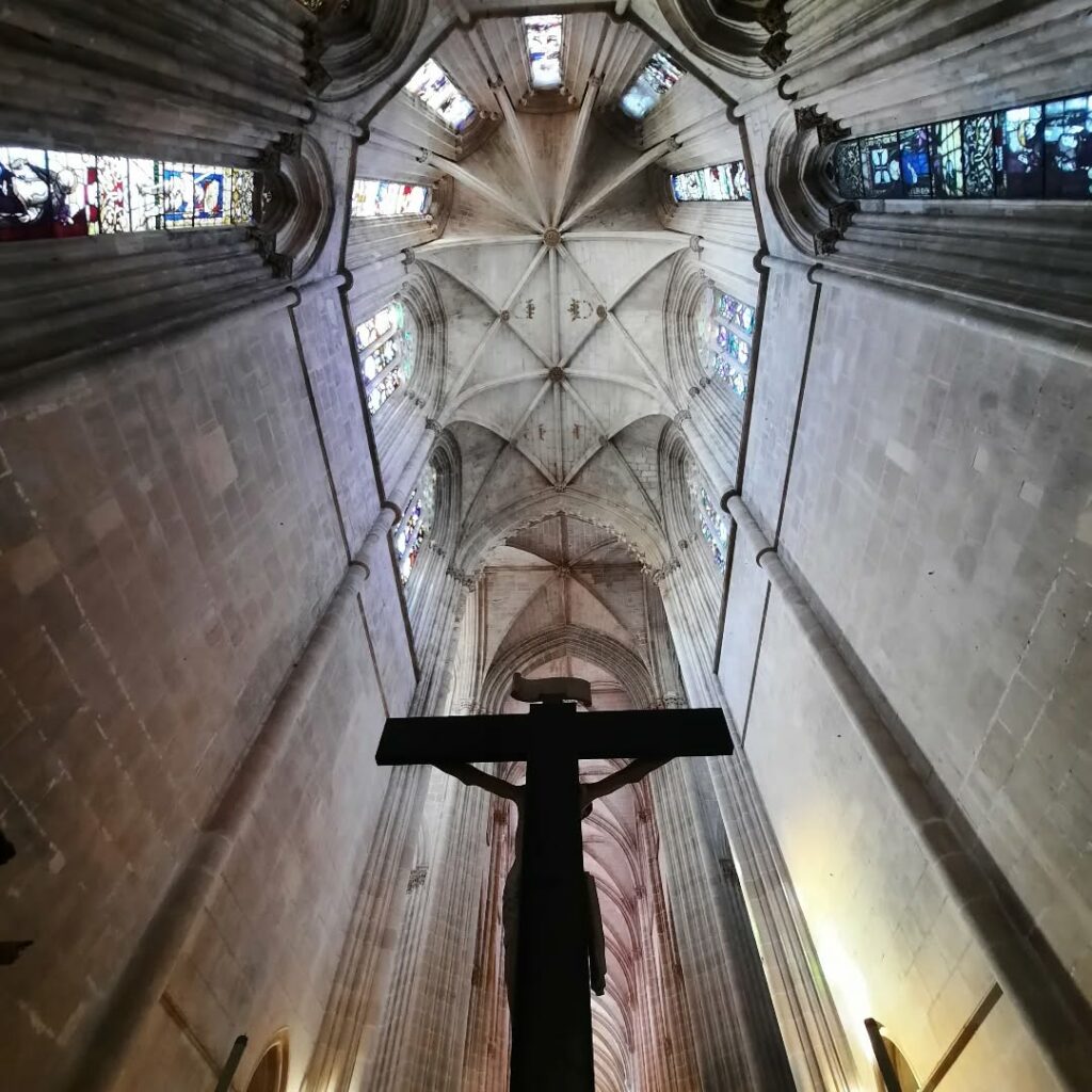 Cruz de Jesus em primeiro plano, com o interior do Mosteiro da Batalha, incluindo a nave e vitrais góticos, ao fundo.