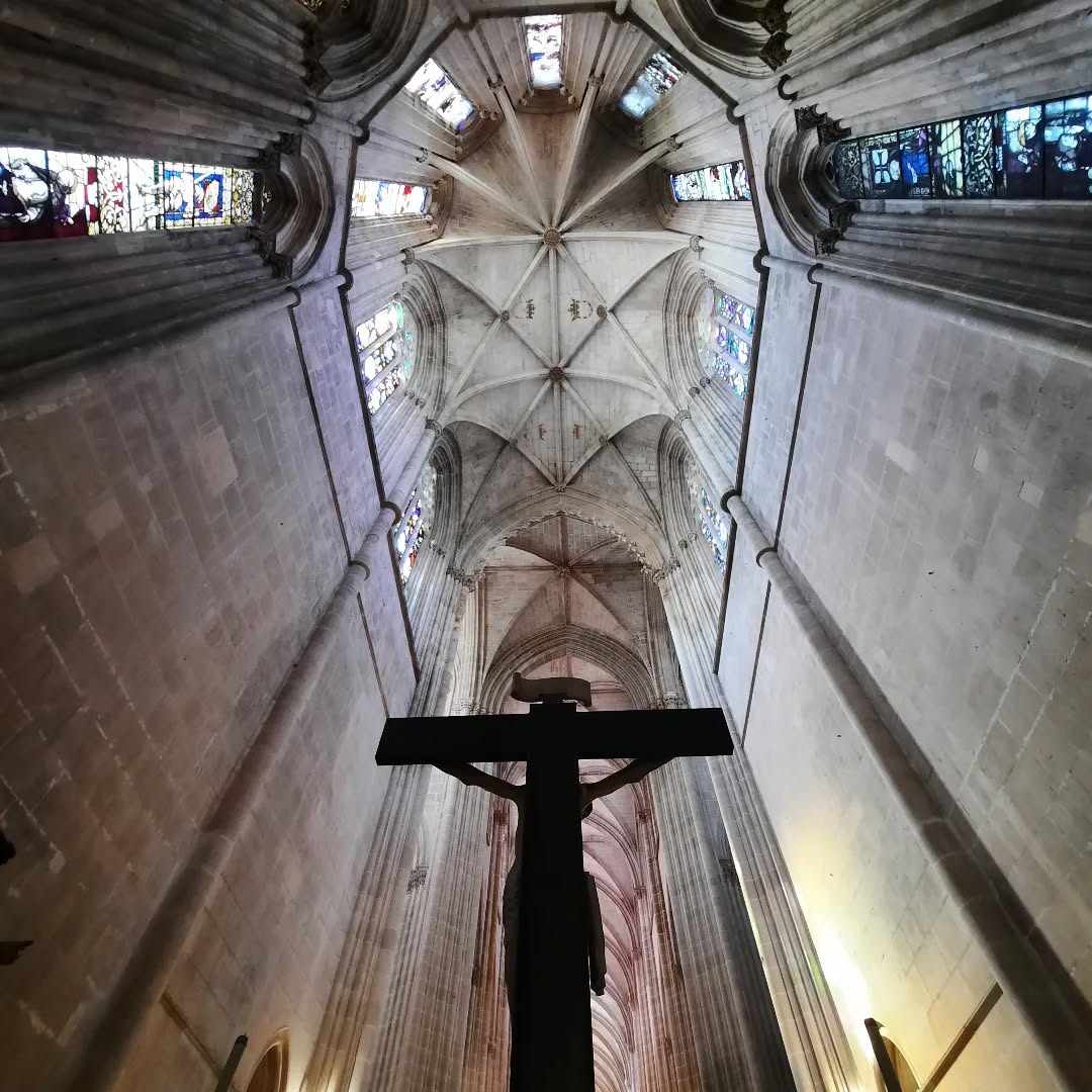 Cruz de Jesus em primeiro plano, com o interior do Mosteiro da Batalha, incluindo a nave e vitrais góticos, ao fundo.