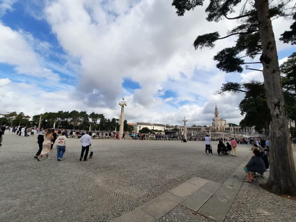 Pessoas caminhando pela Esplanada do Santuário de Fátima sob um céu com nuvens brancas. A Basílica da Santíssima Trindade e a Basílica de Nossa Senhora do Rosário de Fátima estão visíveis ao fundo